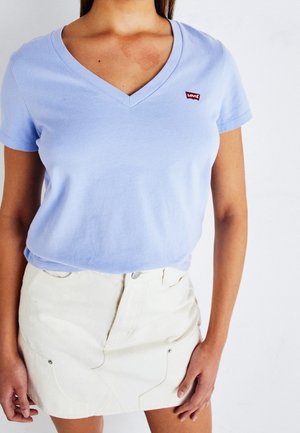 Woman wearing light blue V-neck short-sleeve Levi's T-shirt and off-white high-waist skirt, standing with arms relaxed against white background.