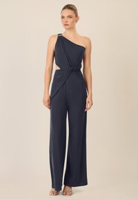 Marineblauwe jumpsuit met éénover ontwerp, geknoopte taille detail en zijkant uitsparingen; gemaakt van soepele, vallende stof; heeft wijde benen.