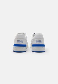 White sneakers with blue accents, featuring a thick sole, smooth leather upper, and "Versace Jeans Couture" logo on the heel.