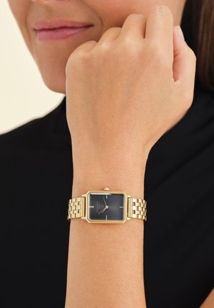 Mujer que lleva un reloj de pulsera rectangular dorado, con esfera negra y diseño minimalista, en su muñeca izquierda, sobre un fondo liso.