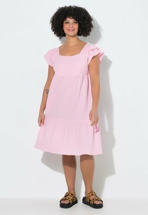 Kvinde med krøllet hår iført en lys rosa knælængde kjole og leopardmønstrede platåsandaler, står mod en hvid baggrund.