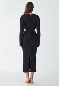 Black long-sleeve dress with cut-out sides, fitted silhouette, and a rear slit. Made of soft fabric, with a subtle texture and back zipper.