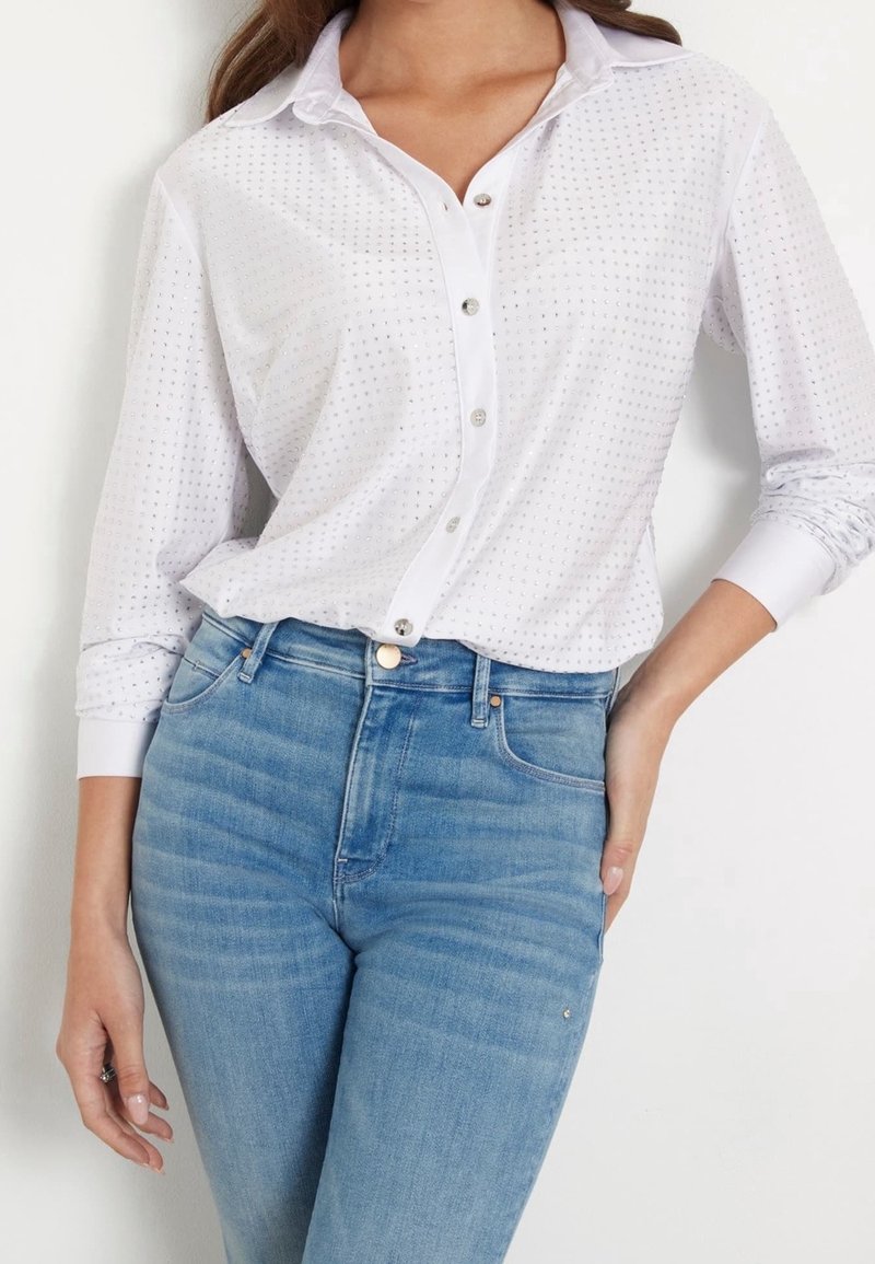 Femme portant une chemise blanche à petits trous et un jean taille haute bleu clair, posant avec une main sur la hanche.