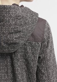 Grey patterned jacket featuring a hood, with a textured fabric and detailed geometric designs on the back. Brown shoulder accents.
