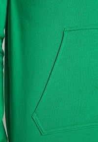 Sudadera verde de tejido suave, con un gran bolsillo frontal y costura en contraste. Textura suave y diseño sencillo.