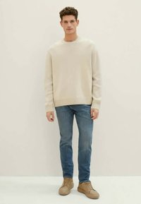 Suéter de punto beige claro, jeans ajustados azul y zapatillas color arena. Diseño simple, textura suave y paleta de colores neutros.