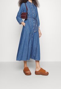 Denim button-up dress with long sleeves, a waist tie, and side pockets. Paired with brown platform slide sandals. Smooth texture and relaxed fit.