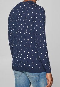 Marineblauer Pullover mit weißen Punkten, Rundhalsausschnitt, langen Ärmeln, geripptem Saum und Bündchen, hergestellt aus einem weichen Stoff. Kombiniert mit hellblauen Jeans.