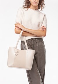 Borsa a tote in pelle beige con un pannello testurizzato, dotata di doppi manici e un tag decorativo. Materiale morbido e liscio con una forma strutturata.