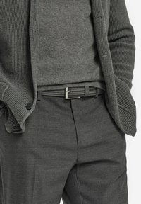 Man in een donkergrijze gebreide cardigan, grijze trui, grijze pantalon en een gevlochten zwarte riem met een zilveren gesp, met zijn handen in zijn zakken.