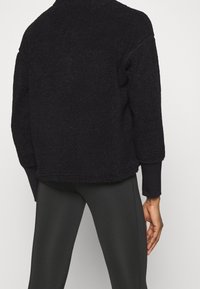 Czarna, futrzana kurtka z polaru o luźnym kroju, z długimi rękawami i okrągłym dekoltem. Materiał ma teksturowany, miękki wygląd.