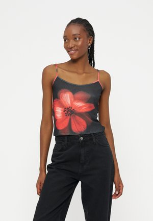 Femme souriante portant un débardeur noir avec un grand motif de fleur rouge et un pantalon noir taille haute, debout devant un fond blanc uni.