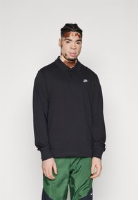 Camisa polo preta de manga longa com colarinho, com um logo Nike branco no peito, usada com calções de desporto verdes.