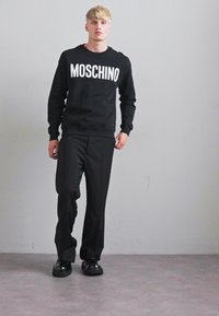 Blond man wearing black Moschino sweatshirt, black trousers, and black shoes, standing against a plain light gray wall.