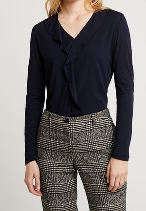 Vrouw draagt een marineblauwe top met lange mouwen en V-hals met ruches aan de voorkant, gecombineerd met zwart-wit geruite high waist broek.