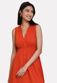Vestido corto naranja con escote en V, diseño sin mangas, cintura fruncida y falda fluida. La tela parece ligera y con textura.