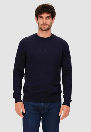 Granatowy sweter z okrągłym dekoltem, długimi rękawami i logo "GUESS" w ciemniejszym odcieniu niebieskiego na klatce piersiowej. Gładka faktura materiału.