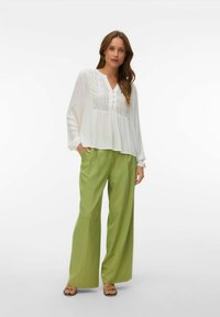 Femme portant un chemisier blanc à manches longues avec des détails en œillets et un pantalon large ample vert clair, debout avec une main dans la poche.