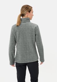 Grijze fleecjacket met hoge kraag en lange mouwen. Heeft een gestructureerd oppervlak en een afgeronde zoom. Draagt samen met zwarte broek, gezien vanaf de achterkant.