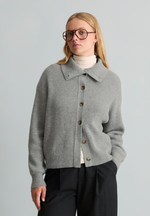 Femme portant de grandes lunettes, un cardigan en tricot gris boutonné sur un col roulé blanc et un pantalon noir, debout devant un fond uni gris clair.