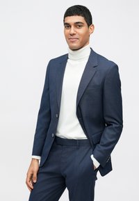 Σκούρο μπλε σατεν κοστούμι με σταθερή εφαρμογή, σακάκι με μονό κουμπί και ταιριαστά παντελόνια. Συνδυασμένο με ελαφρύ πουλόβερ με ψηλό γιακά.