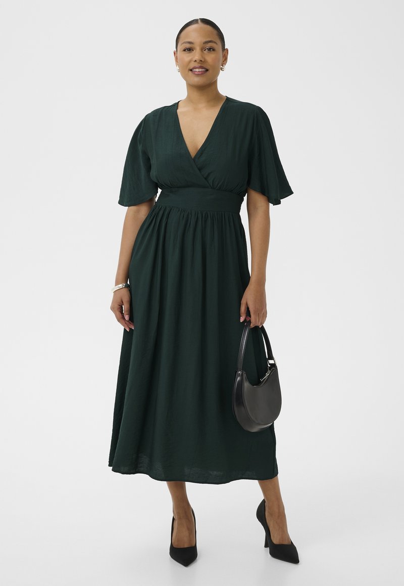 Ženska, oblečena v temno zeleno midi obleko z V-izrezom in rahlo razširjenimi rokavi, drži črno torbico, ima črne pete, stoji pred belim ozadjem.