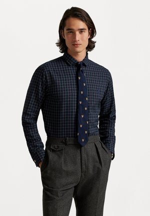 Camisa de cuadros azul marino con líneas verdes y blancas, combinada con una corbata azul marino con patrones de cresta dorada y pantalones grises con pliegues.