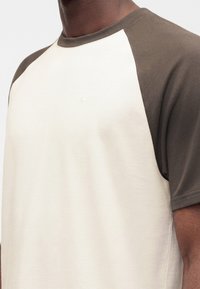 Κοντινό πλάνο ενός ατόμου που φοράει κοντομάνικο, δίχρωμο πουκάμισο με σκούρα ελιά μανίκια και ανοιχτό μπεζ σώμα.