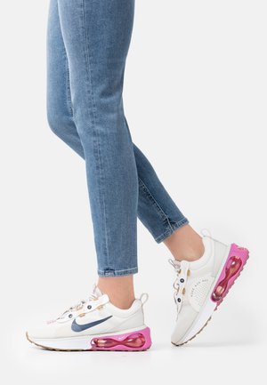 Person wearing light blue jeans and white Nike Air Max sneakers with pink accents, showing side and back views of the shoes.