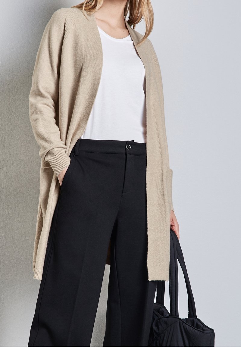 Béžový kardigan s otevřeným předním dílem, žebrovanými manžetami a postranními kapsami, nošený přes bílou košili, doplněný černými kalhotami s širokými nohavicemi a černou taškou.