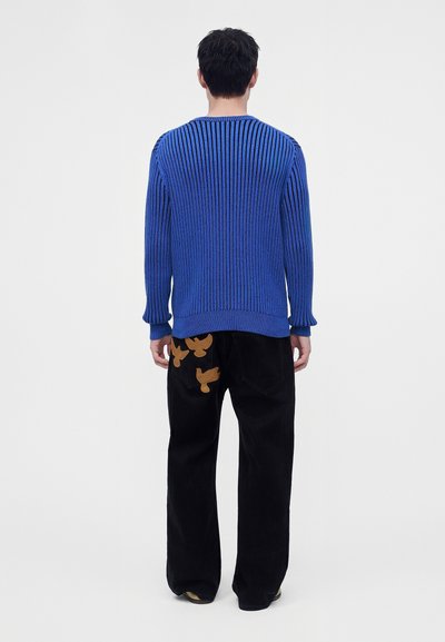 Homme portant un pull côtelé bleu et un pantalon noir avec des patchs oiseaux marron sur la poche arrière, debout devant un fond blanc uni.