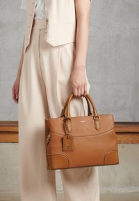 Borsa a mano in pelle marrone con texture, dotata di manici doppi, forma rettangolare e etichetta per bagagli. Presenta cuciture a contrasto e dettagli decorativi.