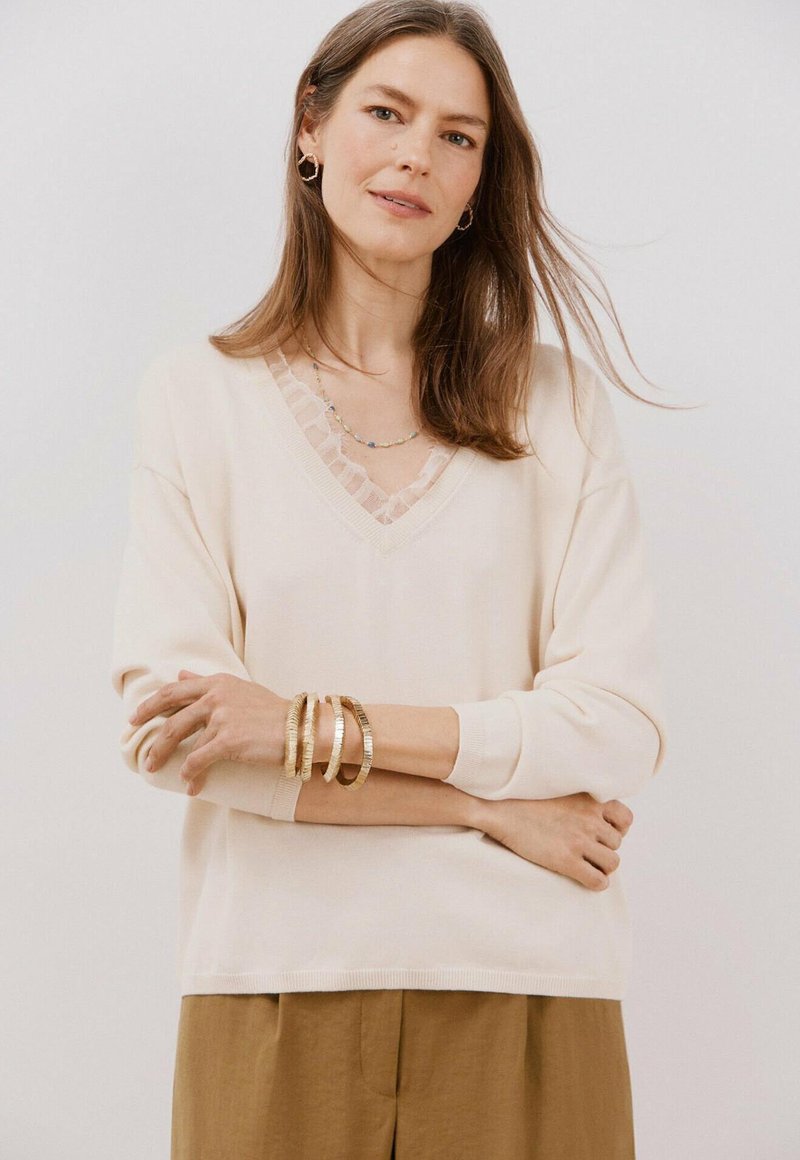 Lichtbeige v-hals trui met textuur kantafwerking, relaxte pasvorm en lange mouwen, gecombineerd met gouden armbanden om de arm van een model.
