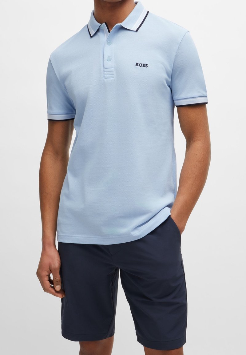 Homme portant un polo bleu clair avec le logo « BOSS » et un short bleu marine foncé, debout avec une main dans la poche sur un fond uni.