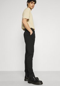 Beige T-Shirt mit kurzen Ärmeln, eng anliegende schwarze Hosen und schwarze Knöchelstiefel mit gelben Nähten. Model posiert seitlich vor einem schlichten Hintergrund.
