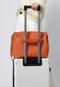 Orange texturerad läderhandväska som vilar på en vit hård resväska, säkrad med ett svart utdragbart handtag för vagn.