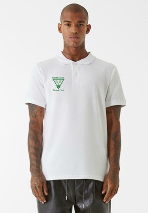 Hvit bomullspolo skjorte med grønn "9NI M SENSE. Tennis Club" logo. Korte ermer, krage, flat strikket tekstur. Brukt med svarte bukser.