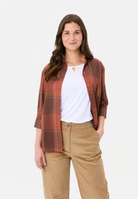 Kariertes Hemd in Rostfarbe mit einer durchgehenden Knopfleiste über einem weißen T-Shirt mit Schlüsselloch-Ausschnitt, kombiniert mit beigen Cordhosen.