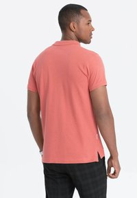 Lyhythihainen korallin värinen polo paita, jossa on teksturoitu kangas, sivuvenytykset ja klassinen kaulus, käytetty ruudullisten housujen kanssa.