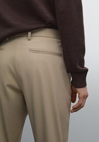 Beige trousers feature a smooth texture, slim fit, and two back pockets with flat closures. The top is a dark, knit sweater with ribbed cuffs.