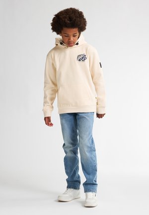 Crème-kleurige hoodie met een voorzak, met een grafische print. Gecombineerd met lichtblauwe jeans en witte sneakers, tegen een neutrale achtergrond.