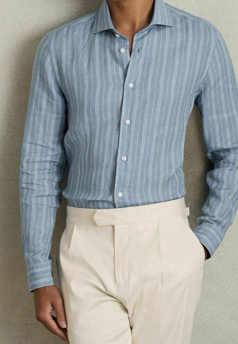 Personne portant une chemise bleu clair à rayures blanches rentrée dans un pantalon beige taille haute avec un détail ceinture.