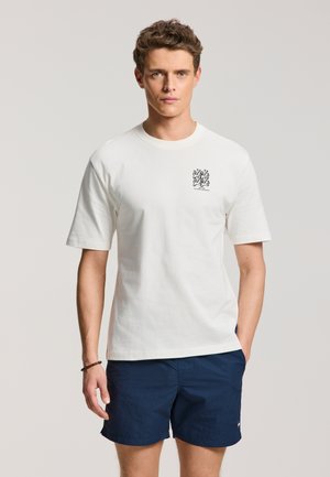 Mand med kort krøllet hår iført en hvid kortærmet T-shirt med lille sort grafik og marineblå shorts, står oprejst.