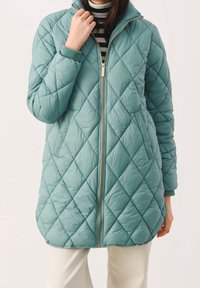 Chaqueta acolchada de color teal con un alto cuello, cierre dorado visible y puños acanalados. Presenta un bordado en forma de diamante y un corte más largo.