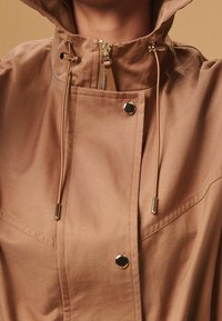 Close-up of beige hooded coat with front zipper, snap buttons, and adjustable drawstring cords with metal tips at the collar.