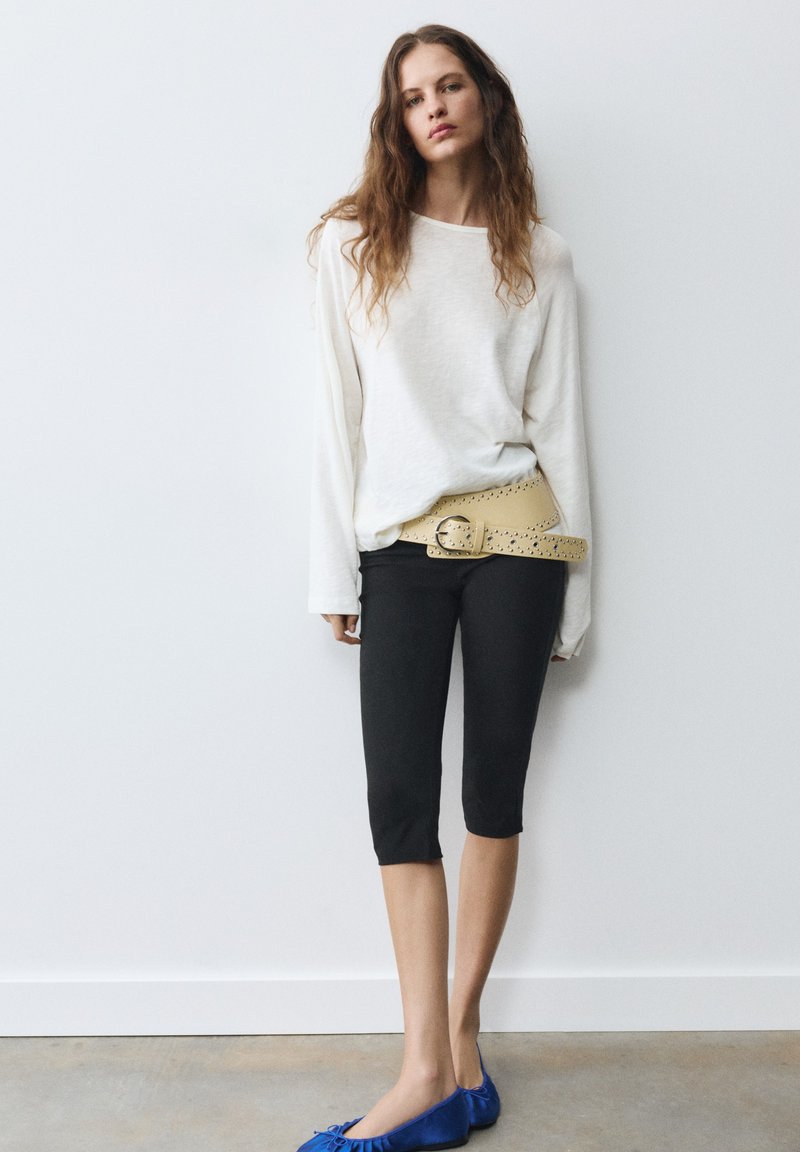 Femme portant une chemise blanche à manches longues, un pantalon court noir, une large ceinture beige cloutée et des ballerines bleues, debout contre un mur blanc.