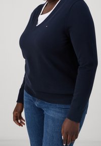 Maglione a scollo a V color blu navy in morbido materiale a maglia, con polsini e orlo a coste. Piccolo logo applicato sul petto. Abbinato a jeans blu.