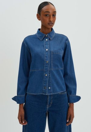 Denim jacket featuring a collared neck, front pocket, button closure, and long sleeves with cuff details. Mid-blue color with a textured finish.