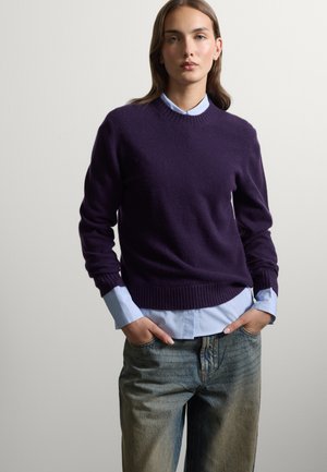 Giovane donna che indossa un maglione viola scuro sopra una camicia azzurro chiaro con bottoni e jeans blu con le mani in tasca, in piedi davanti a uno sfondo semplice.