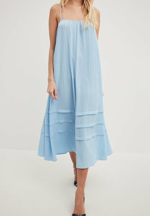 Vrouw draagt een lichtblauwe, mouwloze, gerimpelde midijurk met dunne bandjes en horizontale laagjes, gecombineerd met zwarte hoge hakken.